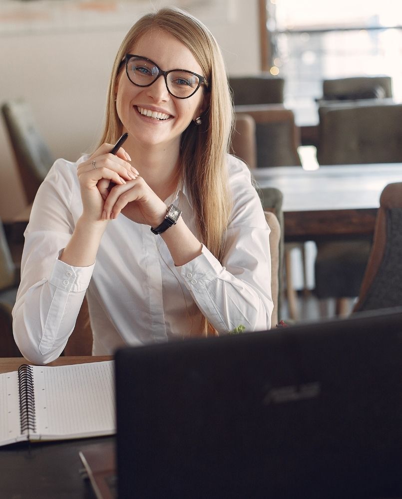 blond woman sits at the desk wearing glasses and smiles at the camera